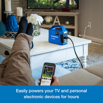 Westinghouse | iGen200s Portable Power Station sits on a table. There are several cords plugged into it. In the foreground someone is holding a phone that is being charged by the power station. A blue banner at the bottom reads, "Easily powers your TV and personal electronic devices for hours".