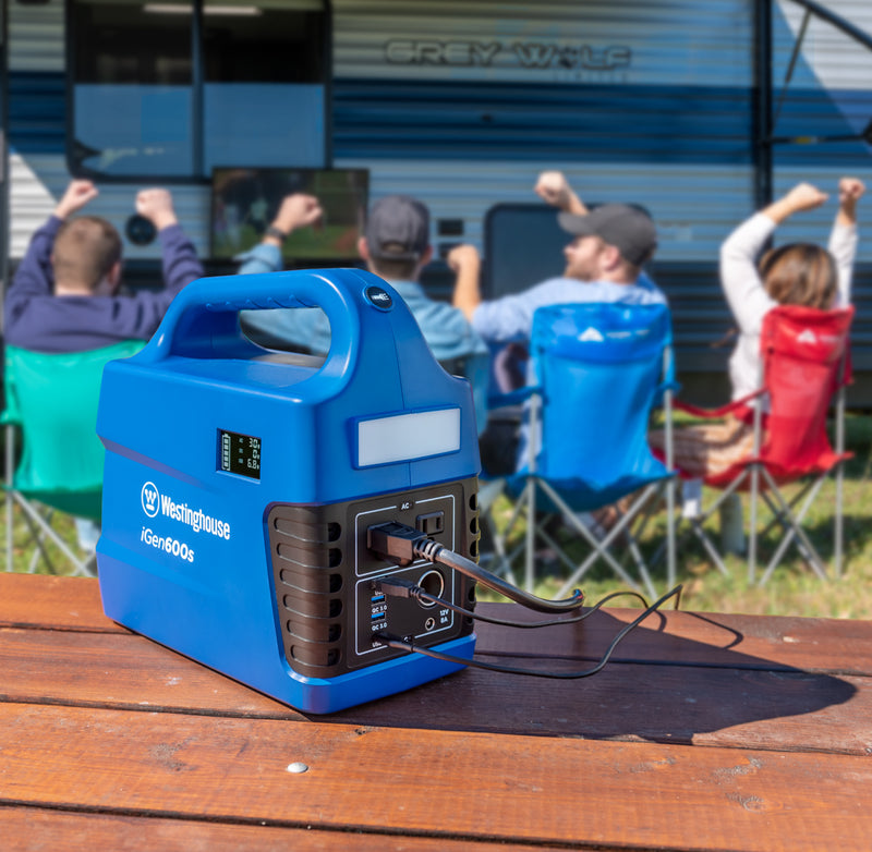 Westinghouse | iGen600s Portable Power Station is shown sitting on a table in the foreground several cables plugged into it. A group of people are watching TV in the background in front of a camper.